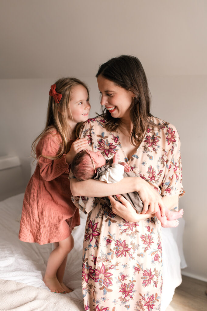 A photo depicting a young brunette mother holding her newborn daughter and smiles at her toddler  in an example of how to select complementary colors in how to plan your family photos wardrobe. 