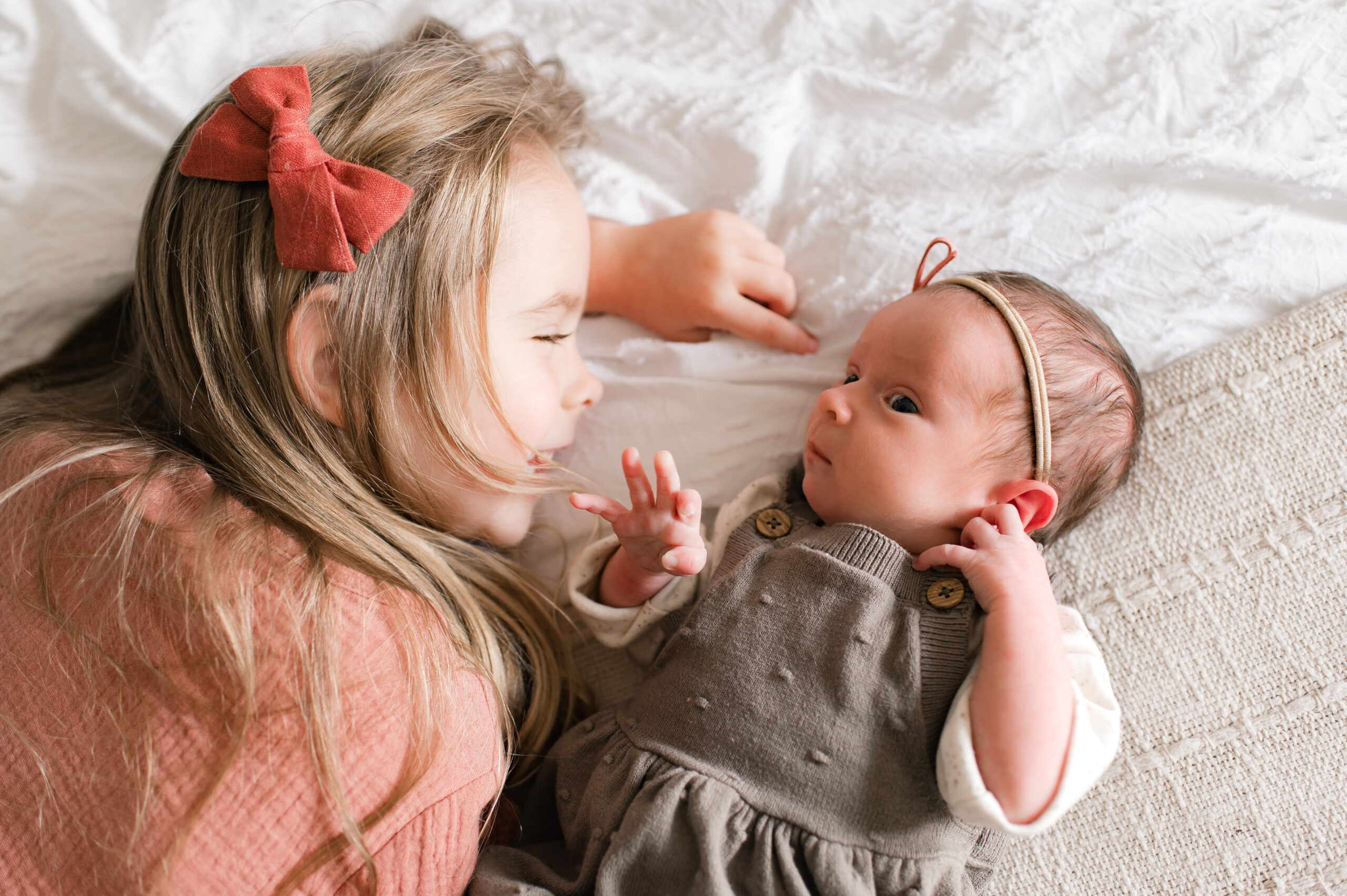 A young girl wearing a burnt orange velvet bow and a light orange linen dress smiles lovingly at her newborn baby sister wearing a brown sweater dress during a family photo session with Mel B Photo, a Cypress TX photographer.