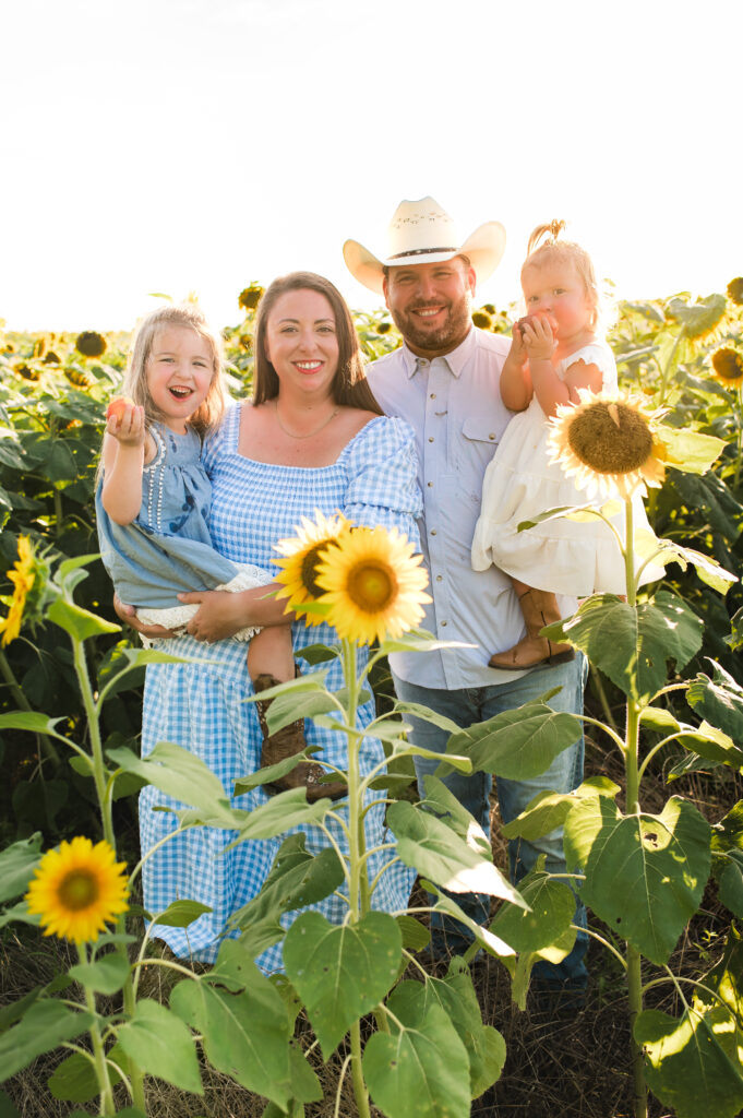 A family of four smiles in a Cypress TX area sunflower field wearing complementary shades of blue in various prints and fabrics.  This image is used as an example of building your family wardrobe around one piece of clothing.