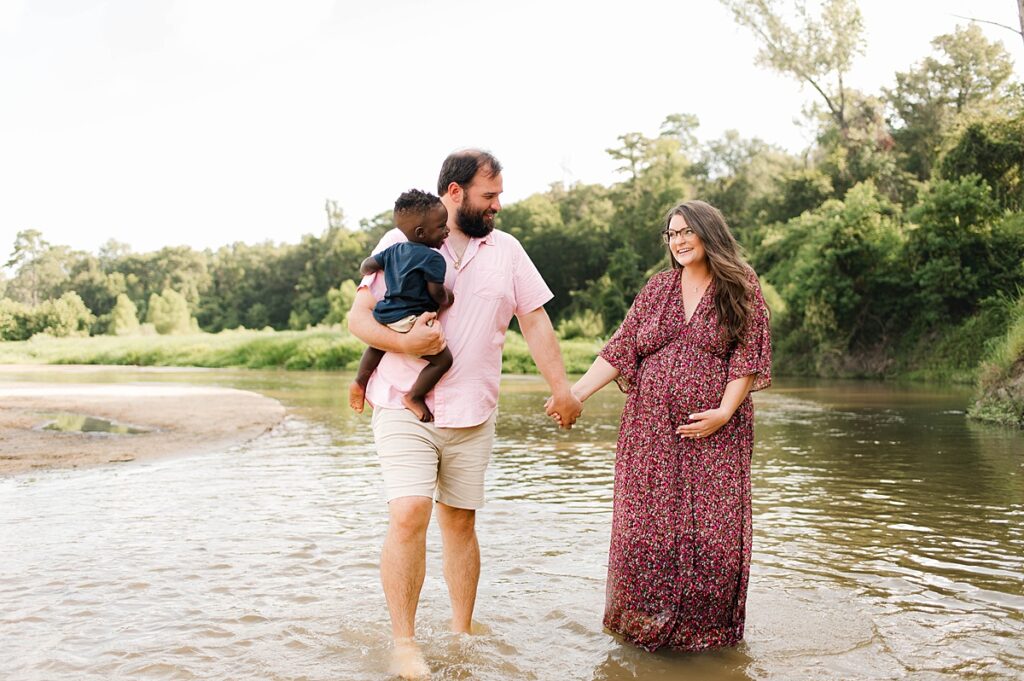 An expectant mother holds hands with her husband who holds their son as they wade through water for family portraits houston tx
