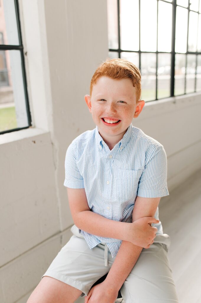 A young boy smiles wearing a light blue seersucker shirt and shorts in a natural light portrait studio in  Houston, Texas 