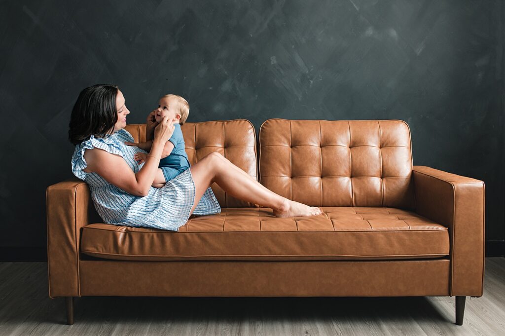 A mother in a blue plaid dress holds her baby on her lap seated on a tan leather couch in a portrait studio in Houston