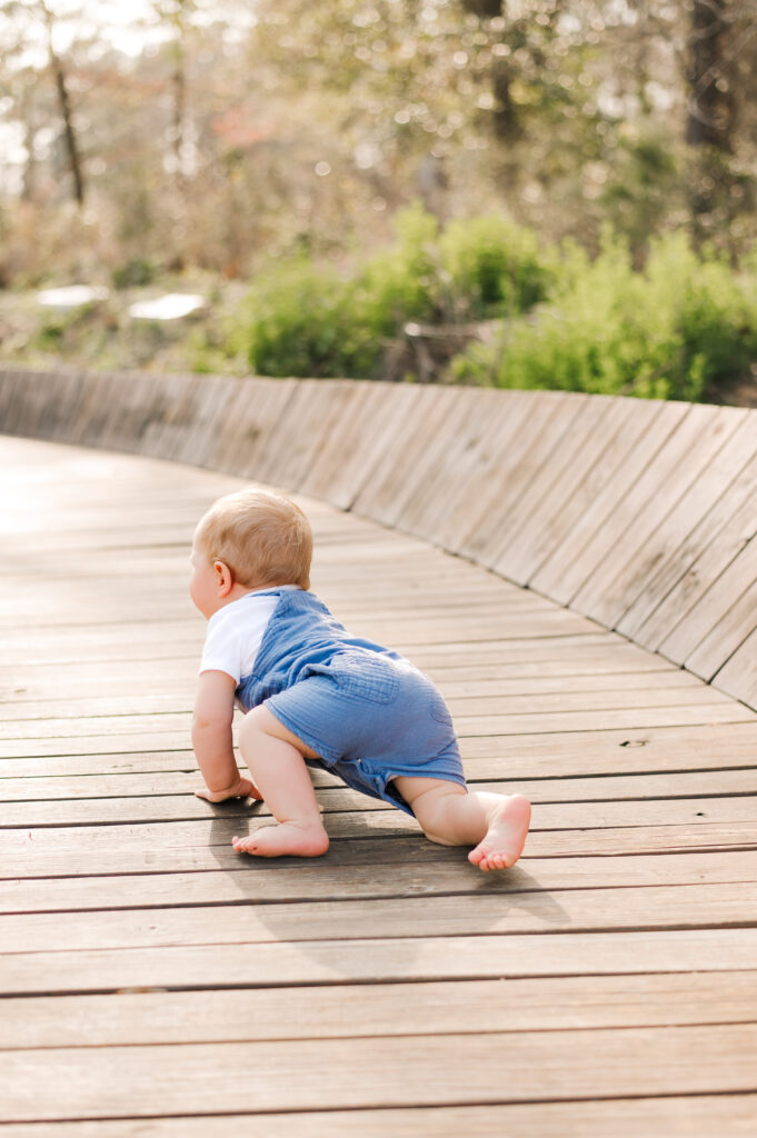 An infant boy wearing blue overalls crawls away from the camera on a wooden bridge in a Houston photo taken at Memorial Park 