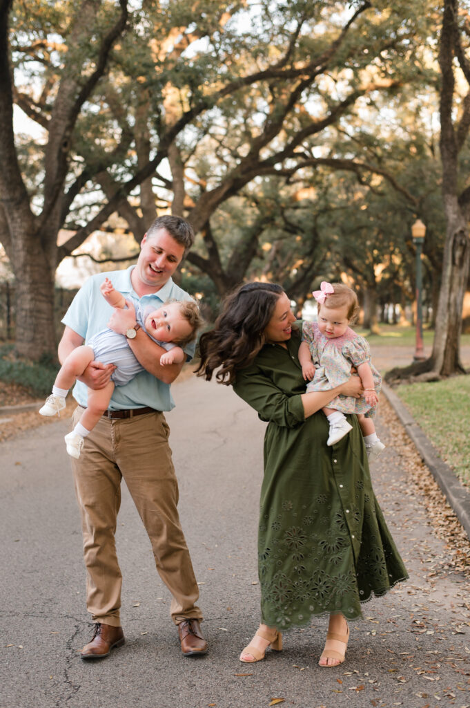 A young couple smile and laugh as they hold their young daughters in family photos taken on North Blvd, one of the best photo locations in Houston 