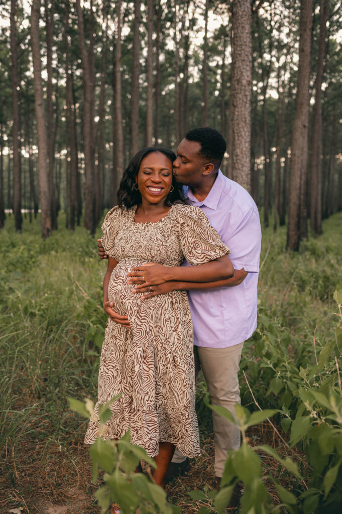 An expectant mother in a flowy dress and her husband rest their hands on her baby bump during a maternity session at WG Jones State Forest 