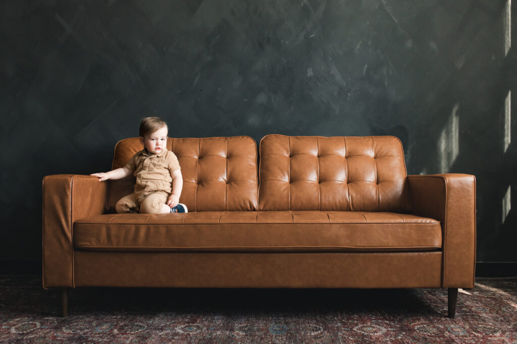 A baby is photographed on a tan leather couch against a charcoal wall at one of  the photo studios in Houston during a family photoshoot with Mel B Photography