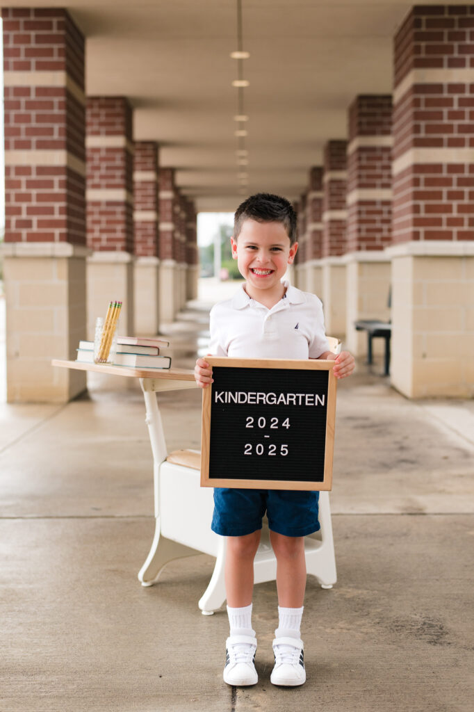 An excited young boy stands smiling before the doors of his new school while holding a letter board in back to school photography taken by Mel B Photo Houston family photographer