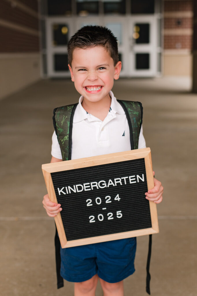 An excited young boy stands smiling before the doors of his new school while holding a letter board in back to school photography taken by Mel B Photo houston family photographer