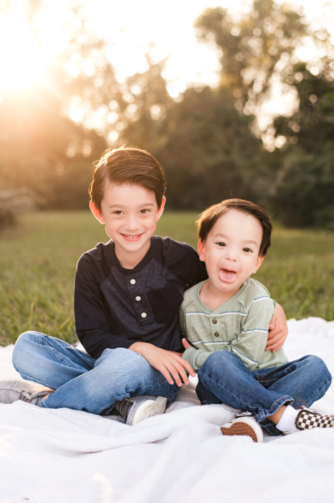 Two young boys wearing jeans smile seated on a blanket with sunlight shining through in the background during family pictures houston