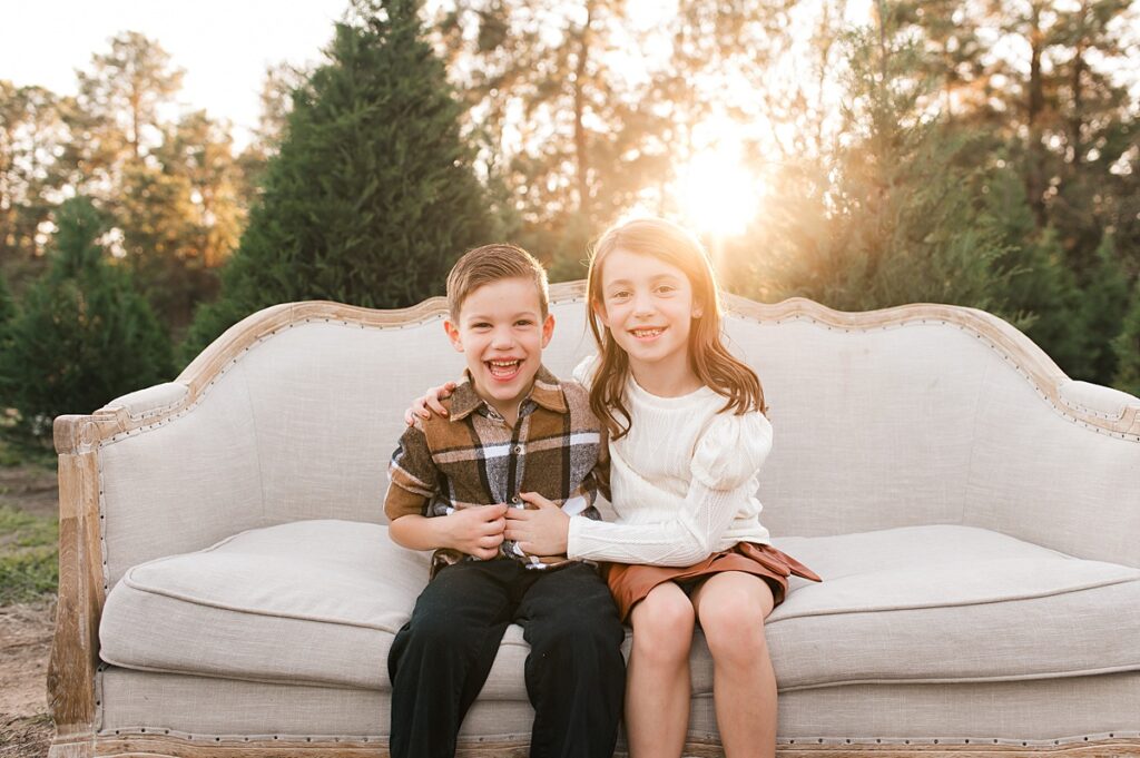 Siblings are seated on a cream colored sofa surrounded by evergreens at a family Christmas photoshoot at Spring Creek Growers in Magnolia, TX