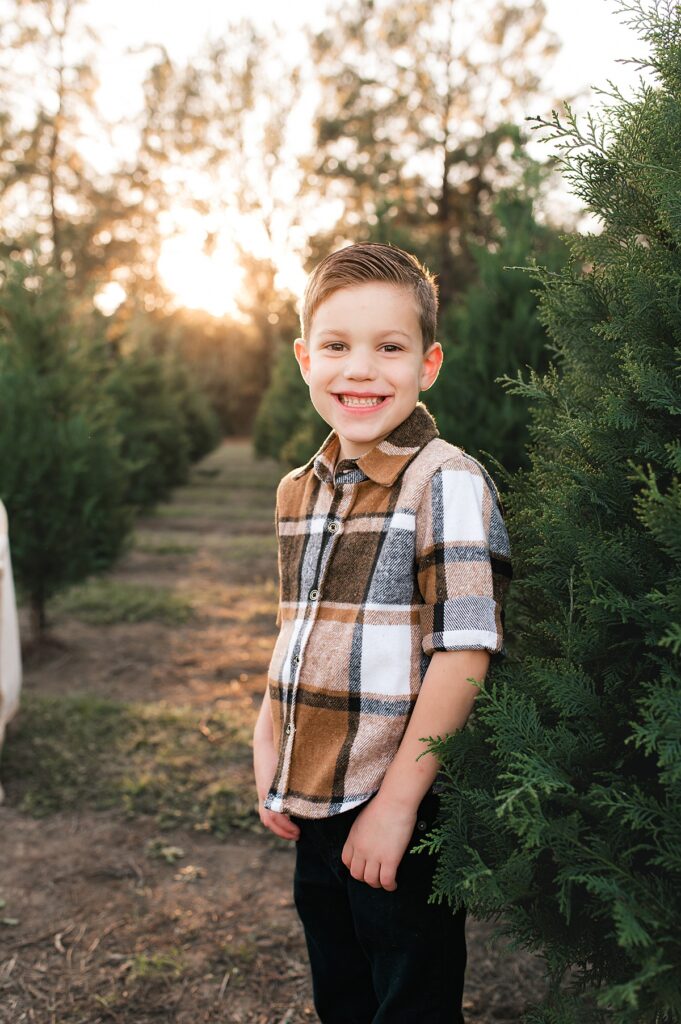 A young boy in a flannel shirt smiles in a row of trees at a Christmas Tree Farm Houston family session