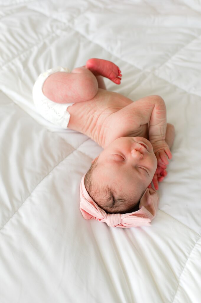 A newborn baby girl wearing a pink bow stretches atop a white comforter during an in-home newborn session with Cypress newborn photographer, Mel B Photo