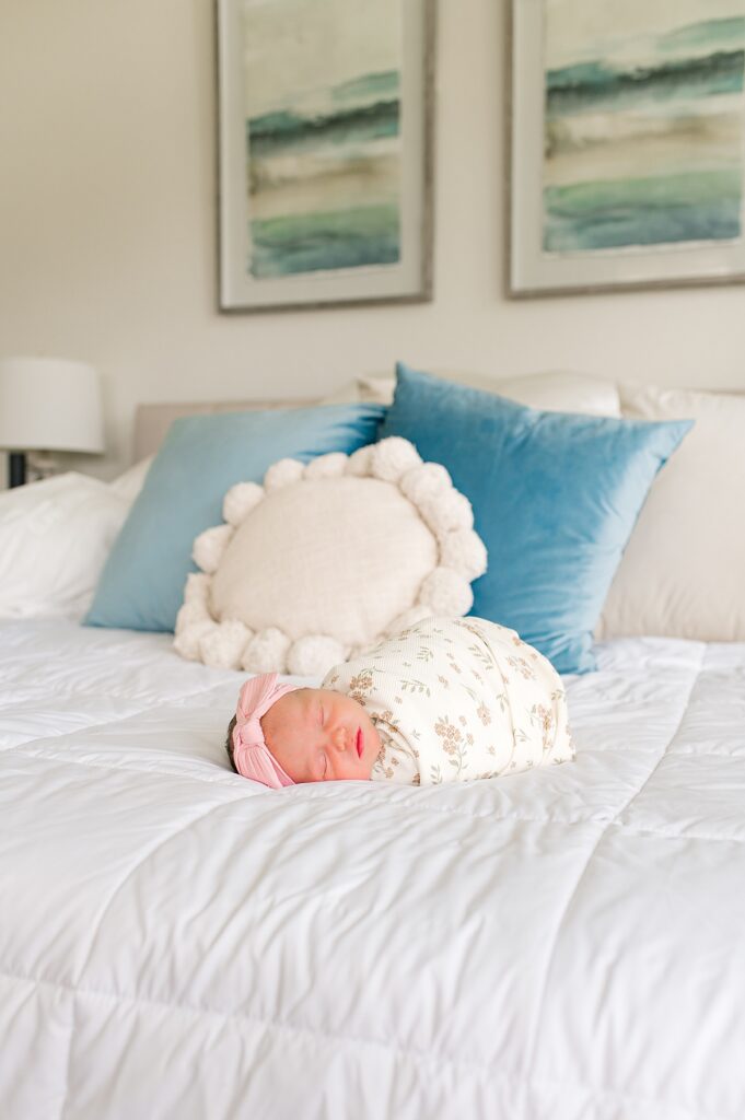 A newborn baby girl wearing a pink headband and a floral swaddle is photographed on a white comforter in the home she shares with her Houston sleep consultant mother & family 