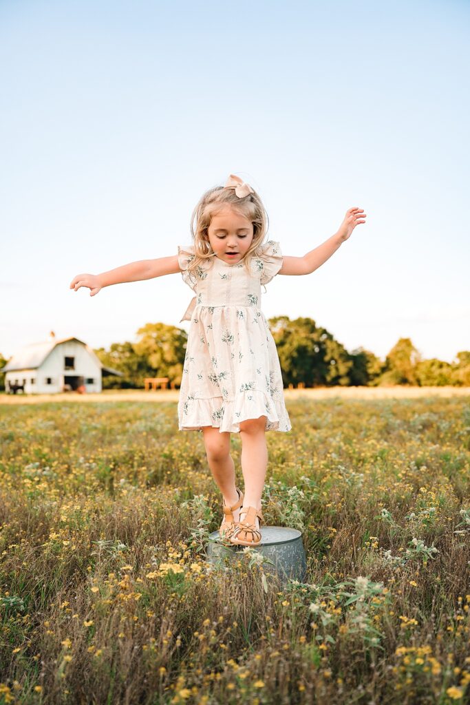 A young girl wearing a floral dress and a bow balances atop a tin bucket at a farm during Houston family photos 