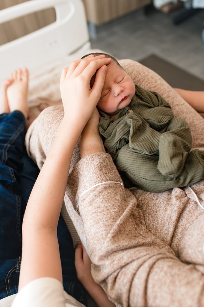 A newborn boy wears an olive green sleeper on his mother's lap in a photo taken during a fresh 48 photography session