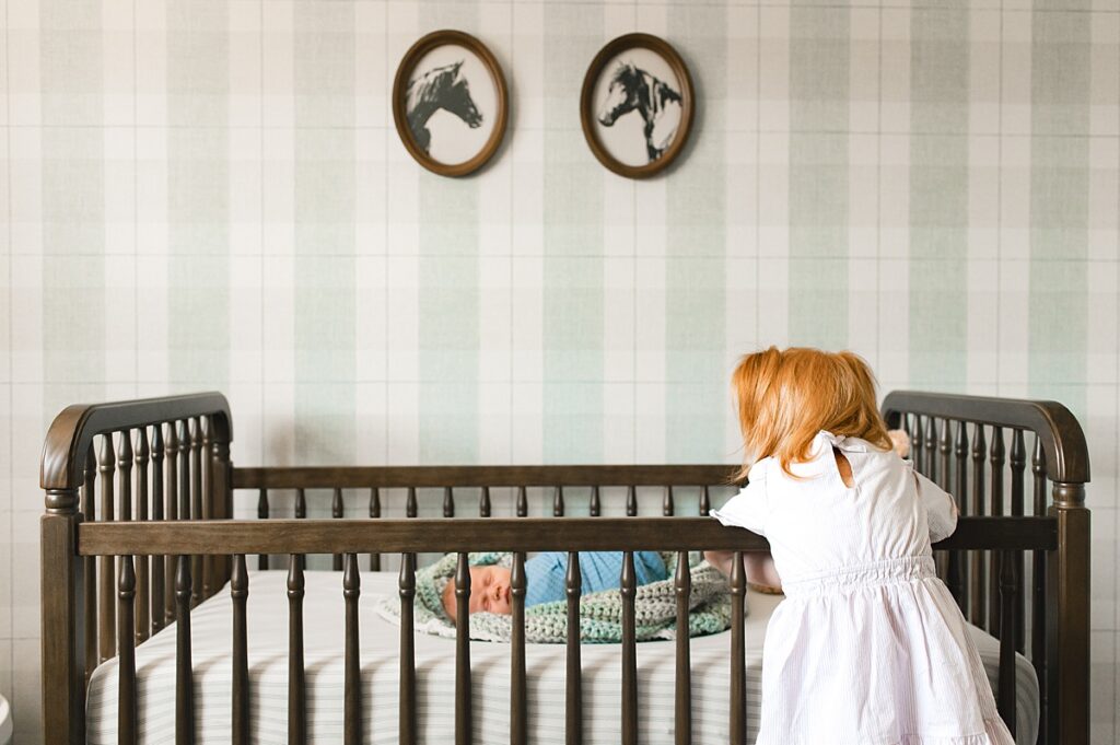 A young girl looks over the side of her newborn brother's crib during Houston newborn nursery photos