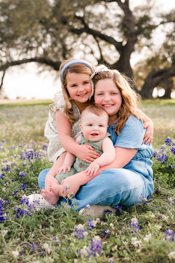 Three young children smile seated on the ground surrounded by Brenham TX Bluebonnets