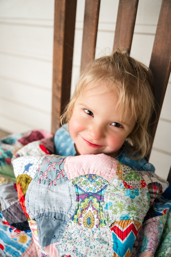 A young blonde girl smiles in a rocking chair while wrapped in a family heirloom quilt in a photo taken for a guide to family fun things to do in houston tx. 