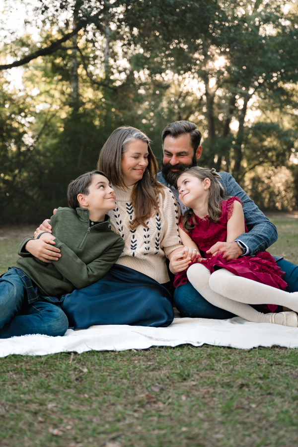 A family of four is seated on a white blanket in a city park houston texas. 