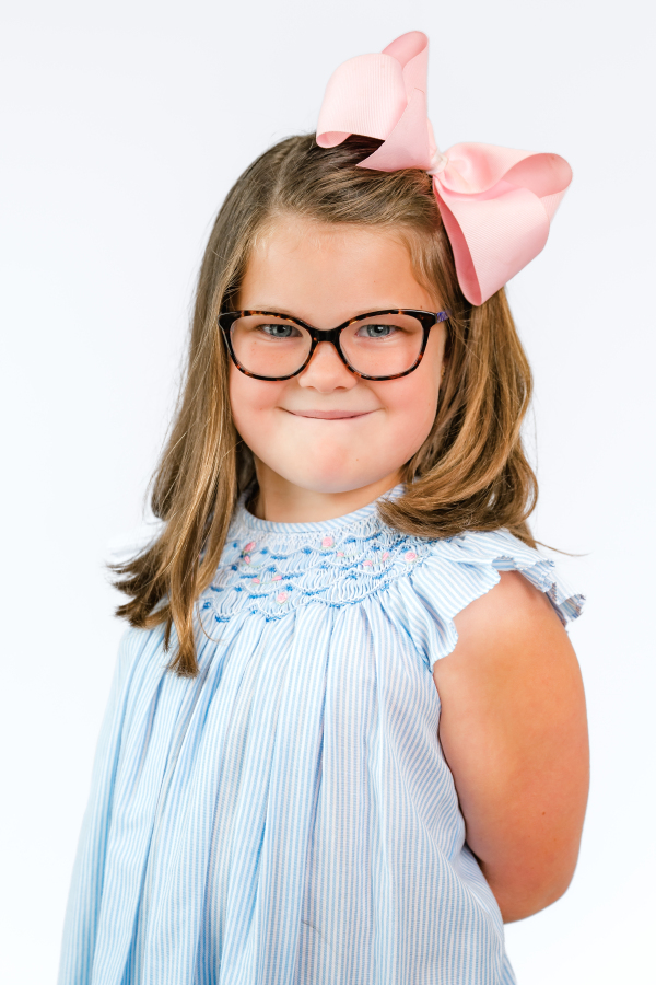 An elementary aged girl wearing a pink bow & glasses smiles in a portrait taken by a Houston school photographer.