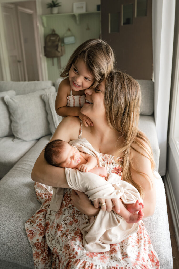 A toddler girl snuggles into her mother, holding a newborn baby in a favorite newborn pose for photography sessions in the home. 