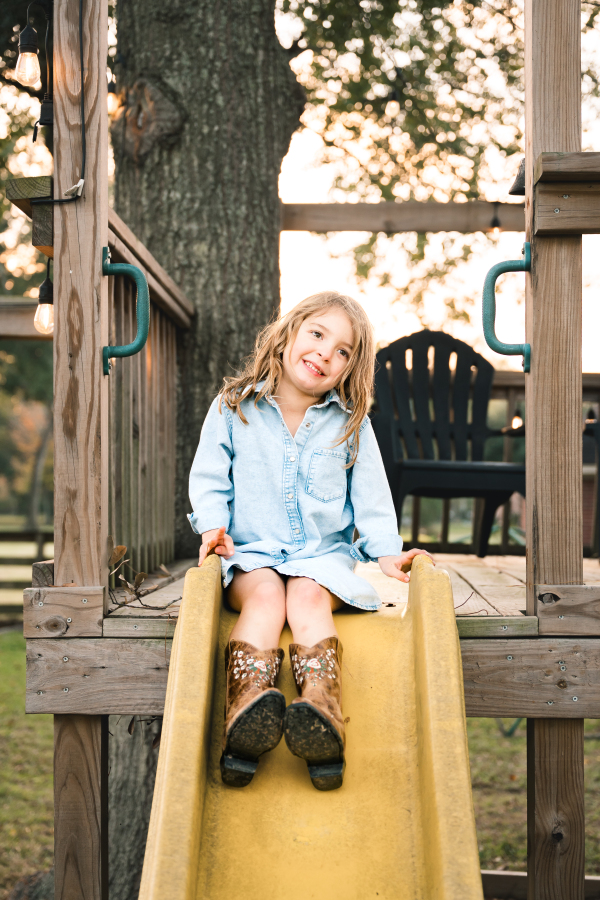 A young girl in a denim dress and cowgirl boots smiles atop her slide in a photo taken by a Houston lifestyle photographer.