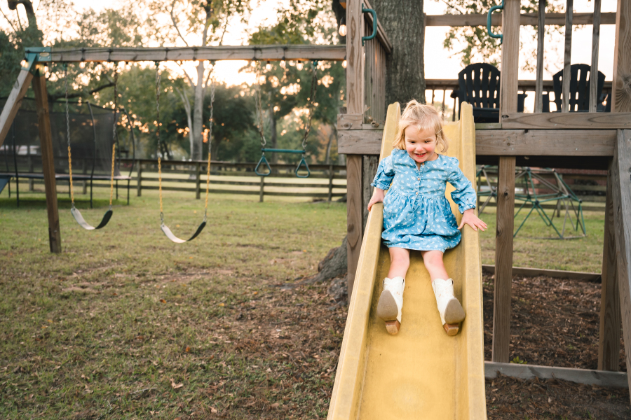 A young girl in a denim dress smiles as she plays on her backyard slide in a photo taken during an in-home family photoshoot in Houston TX.