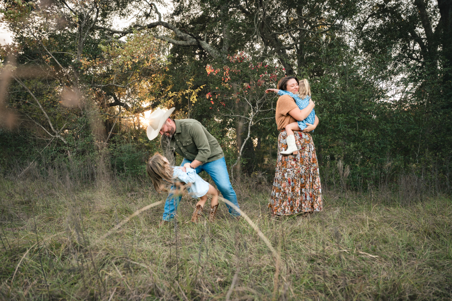 A family of four smile and play in an open field near their home during an at home photoshoot with a Houston lifestyle photographer.