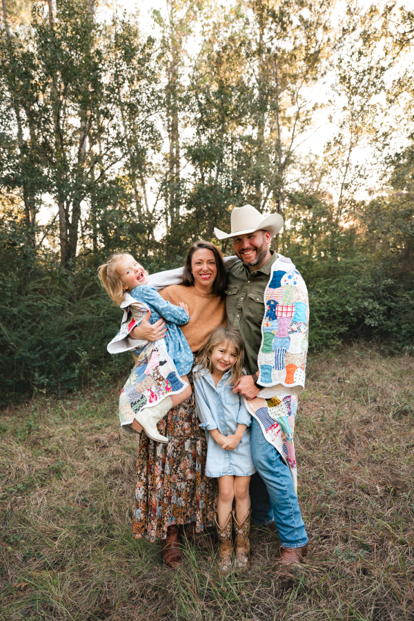 A family of four smiles wrapped in an heirloom quilt during a lifestyle family photo session in Houston TX.
