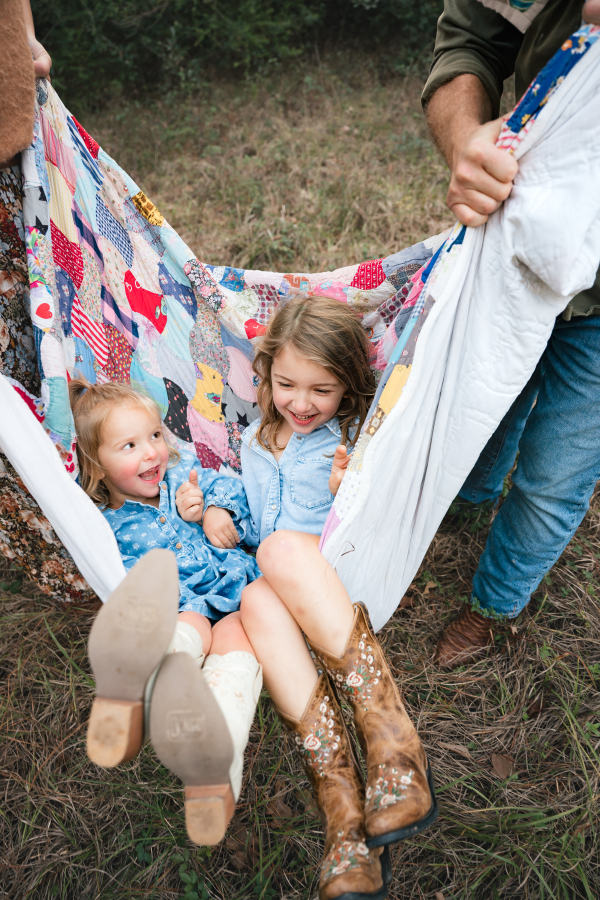 Two young girls smile as their parents swing them on a quilt during a Houston family photo session at their home.