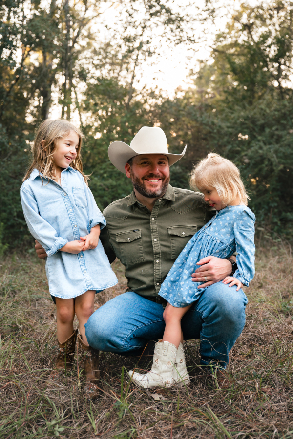 A father wearing a green shirt and cowboy hat poses with his two young daughters on his knees during a session with a Houston lifestyle photographer.